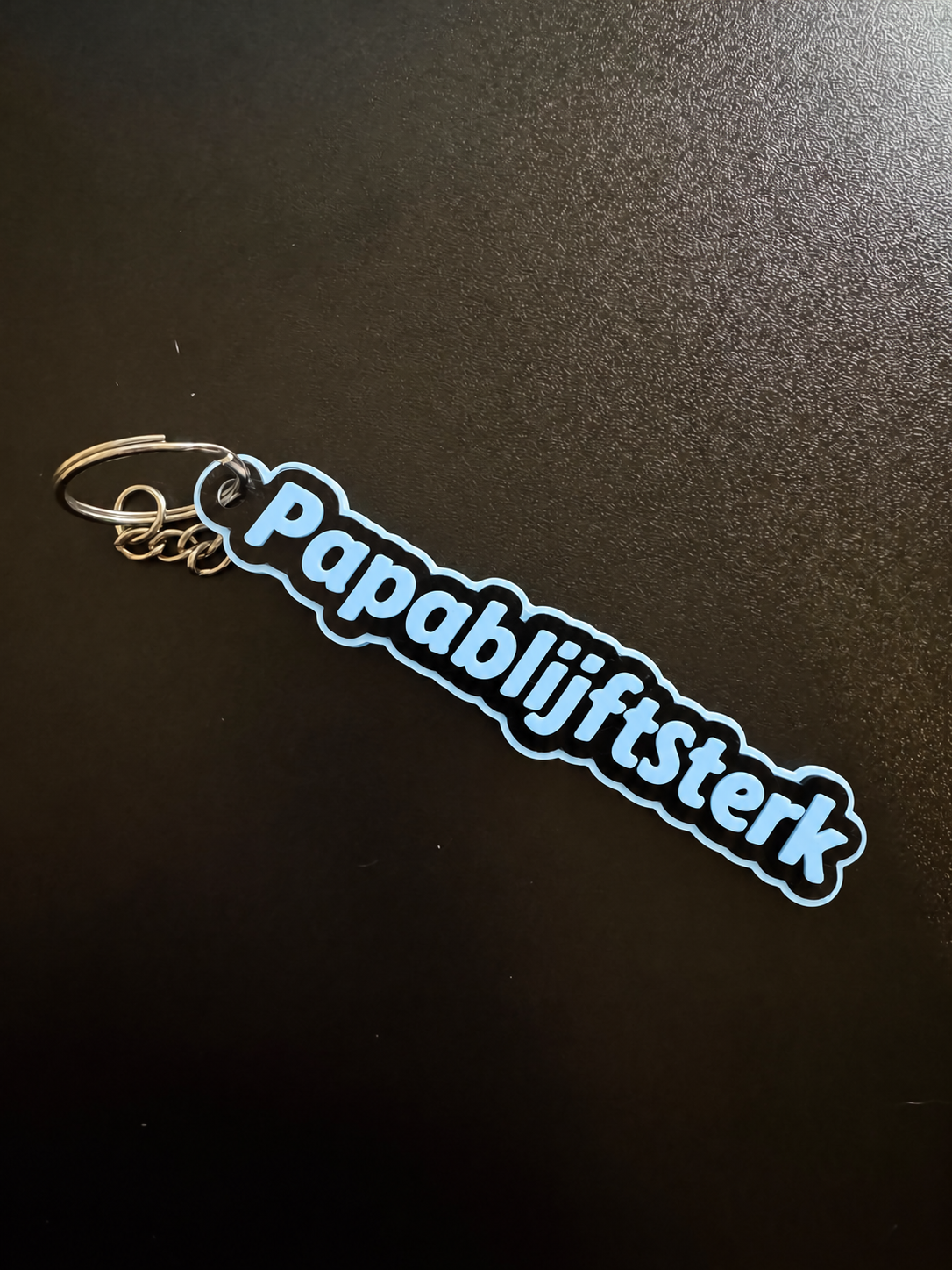 Originele foto van de platte zwarte PapaBlijftSterk-sleutelhanger met babyblauwe letters en rand, metalen ring en kettinkje.